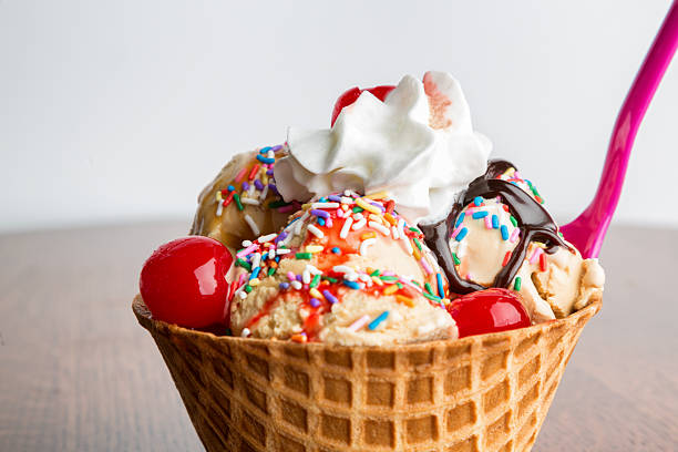 Fancy Ice Cream Sundae with Hot Fudge, Sprinkles, Cherries Waffle Cone filled with Multi-flavored ice cream with Hot fudge, an cherry sauce with whipped  cream and sprinkles and maraschino cherries on an oak table with a pink plastic spoon in the side. ice cream cone photos stock pictures, royalty-free photos & images