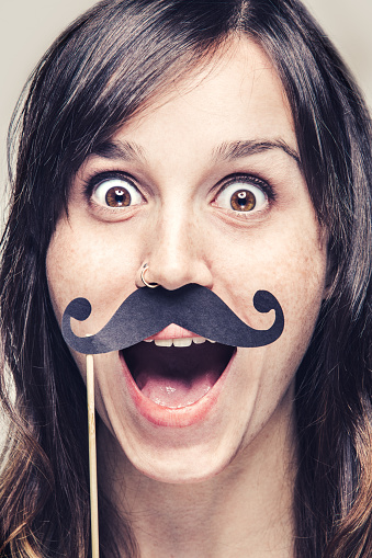 A portrait of a young woman holding up paper mustache on a stick to her face with a huge open mouth smile and big eyes.