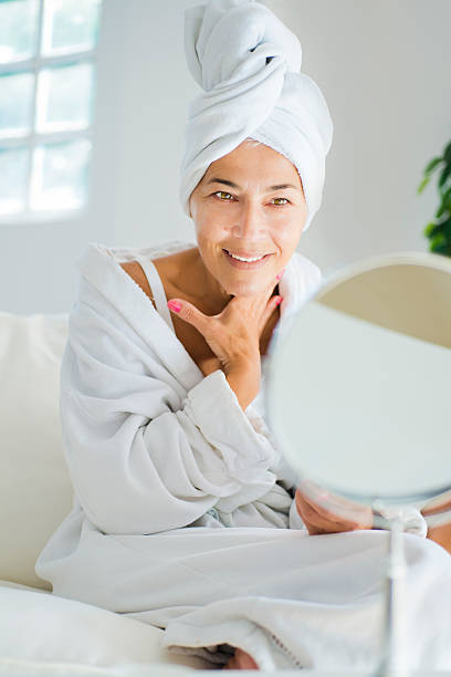 Mature woman and body care Beautiful mature woman applying a cream on her face after showering looking in mirror stock pictures, royalty-free photos & images