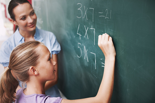 A young girl doing maths on the board as her teacher watches smiling