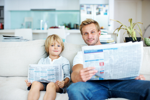 Cute little boy pretending to read a newspaper like his father while on the sofa at home