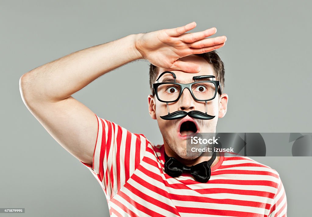 Shocked man wearing fake mask Portrait of shocked young man wearing funny mask staring at the camera with mouth open. Studio shot, gray background. Young Men Stock Photo