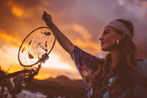 Boho style girl holding up a dream catcher and looking away wistfully on a summer evening