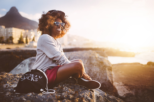 Happy hipster teen girl smiling and sitting by herself on some rocks at the beach