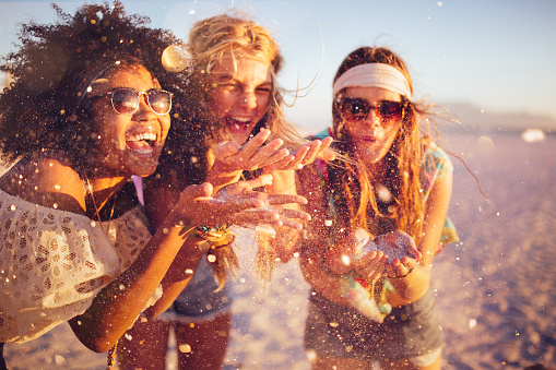 Mixed race group of girls blowing colourful confetti from their hands happily on a beach at sunset