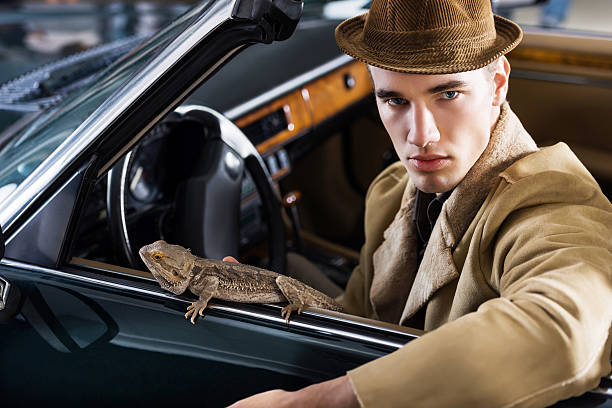 Young fashion retro male model wearing a hat sitting in a car with iguana and looking at the camera.