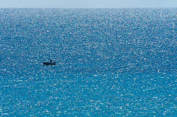 Lone fisherman in small boat on rippled blue-green sea with white reflections of low sun on choppy wavelts, narrow band of sky