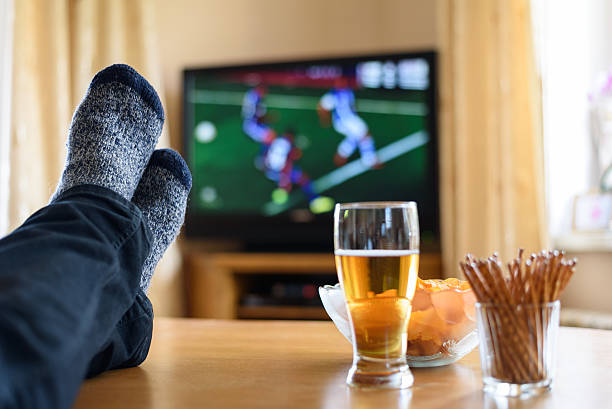 Man watching tv with feet up on table with beer and snacks Television, TV watching (football match) with feet on table and huge amounts of snacks - stock photo football socks stock pictures, royalty-free photos & images