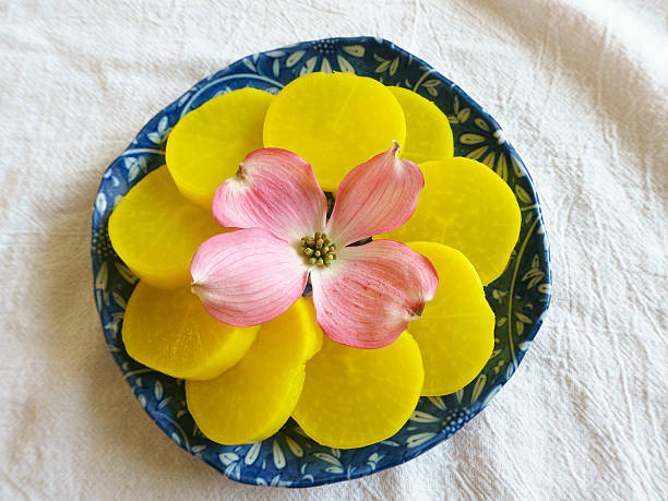 Pickled Daikon Radish This picture depicts a yellow, pickled daikon radish with a decorative dogwood flower in the center of a blue plate.  This banchan, vegetable dish sits on a white, textured background and was taken with a mirrorless camera. banchan stock pictures, royalty-free photos & images