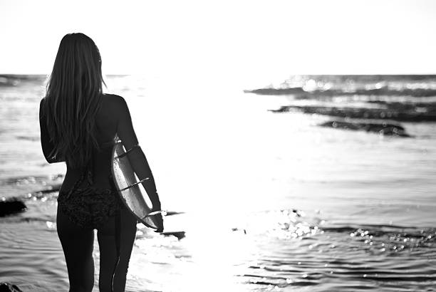Black and white silhouette of blonde surfer girl holding her surfboard and looking out over the water.