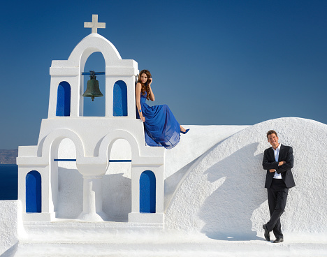 Happy Groom with his beautiful Bride in a blue dress sitting by a typical blue church bell on a rooftop in Oia on Santorini, Greece.
