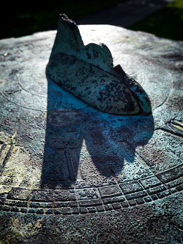 Close-up of a backlit old copper sundial with a broken gnomon, although it's still possible to see where the shadow points when the sun is low, as it is here. Contrasty, low-key image to bring out the texture of the verdigris-covered copper. Sharp focus is on the dial in the foreground.