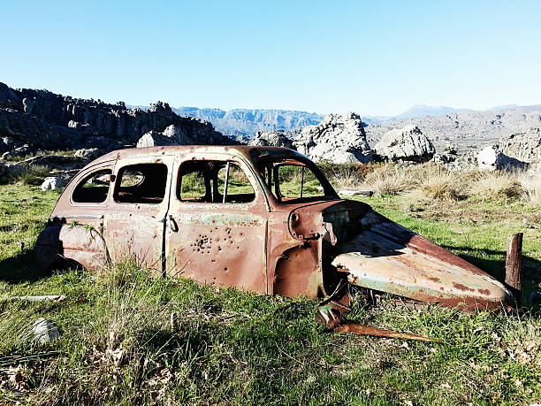 A rusted, abandoned vehicle sits amidst wild overgrown vegetation in front of a remote, white-walled house. The scene evokes decay, neglect, and the passage of time in a desolate landscape.