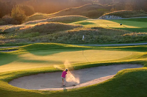 A young junior golfer blasts a ball out of a bunker on a beautiful golf course. Sand is backlit to provide drama. Themes of this image include junior golf, links golf, evening, kids, children, fun, playing, swinging, short game, bunker shot, unrecognizable golfer, Calgary, alberta, Canada, scenic, boys, golf camp, lessons, one person, and lifestyle. 