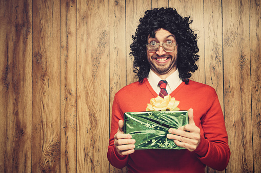 A nerd man with long curly black hair and 1980's retro style stands in front of a wood paneled wall holding a Christmas gift, an excited, goofy expression on his face.  He wears a Christmas colored red and green tie and sweater.  Horizontal with copy space.
