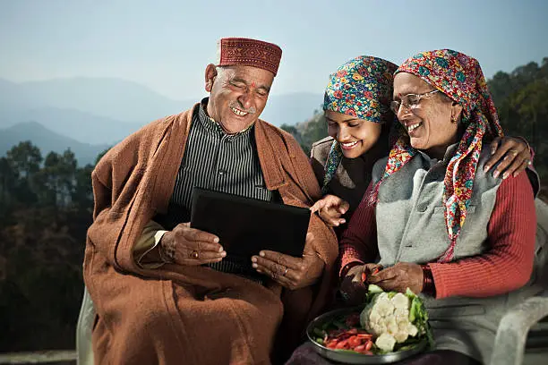 People of Himachal Pradesh:  A happy senior man showing laptop to his daughter and wife while his wife chopping vegetables and daughter leaning over her shoulder and pointing something on laptop screen with toothy smile, they are sitting on chairs against natural background of mountains and trees in their traditional dress of Himachal Pradesh, India.