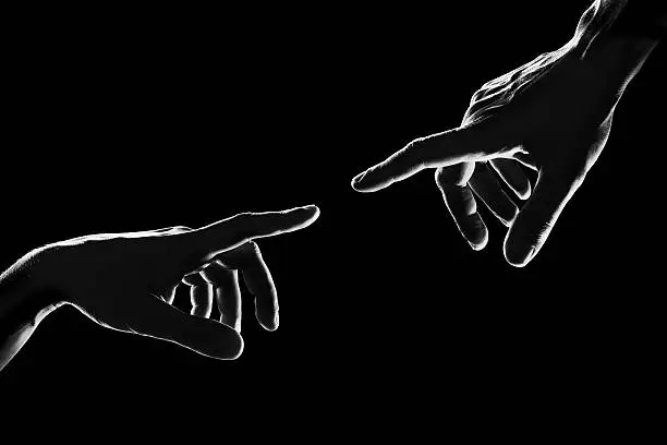 Close up of two hands, reaching each other like Michelangelo's painting on black background. Black and white image, strong contrast, back lit.