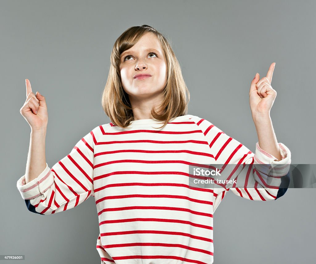 Cute girl pointing Portrait of cute girl wearing striped blouse and jeans, standing against grey background and pointing with index fingers, looking up. Girls Stock Photo