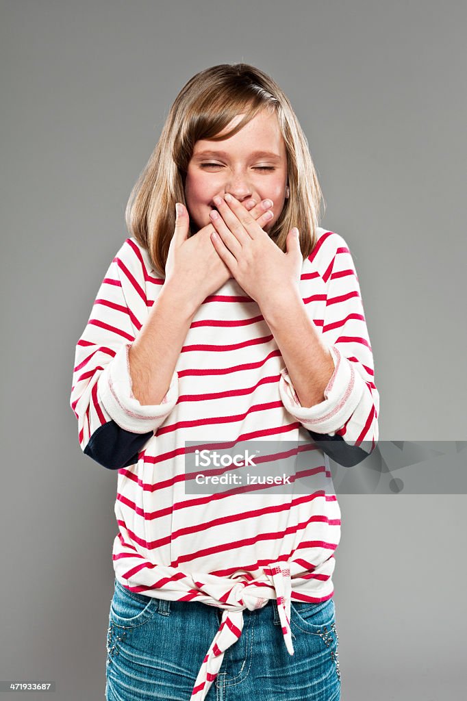 Laugh Portrait of cute girl wearing striped blouse giggling with hands covering her mouth. Studio shot, grey background. 10-11 Years Stock Photo