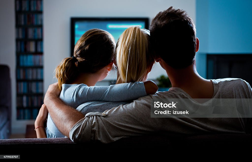 Family watching TV Back view of family sitting on sofa at home and watching TV. Dark tones. Television Industry Stock Photo