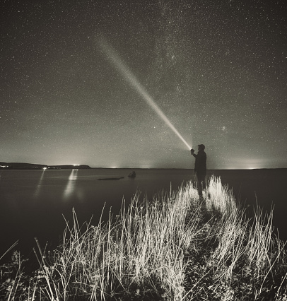 A man stands on a narrow point with a spotlight pointed at Earth's axis which lies very close to Polaris, the North Star.  Long exposure with light painting, toned black and white.