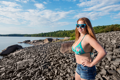This is a horizontal, color photograph of an 18 year old young woman at a lake in upstate New York. She stands on the rocky beach in the Catskill Mountains enjoying a sunny day in a bikini top and sunglasses.
