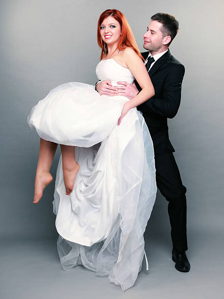 Wedding day. Portrait of happy married couple groom holding red haired bride up in his hands full length studio shot on gray background