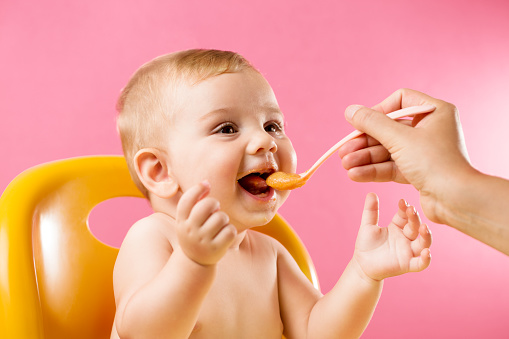 Mother's hand feeding a laughing baby in front of a vivid pink background.