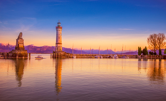Boat passing the famous harbour entrance with the landmark Bavarian lion and lighthouse of Lindau (Bodensee) at sunset. The snow-covered glowing alps in the distance, 