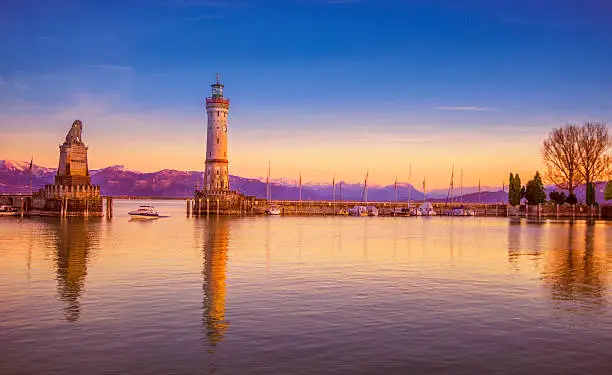 Boat passing the famous harbour entrance with the landmark Bavarian lion and lighthouse of Lindau (Bodensee) at sunset. The snow-covered glowing alps in the distance, 