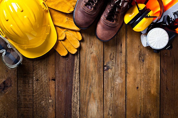 Personal safety workwear border on rustic wood background Personal safety workwear items shot directly above arranged as a border at top of the frame, from left to right are goggles, helmet, gloves, steel toe shoes, earmuff a respiratory mask and a safety vest leaving a useful copy space in the center and bottom. The background is a rustic wood table scratched and stained with vertical stripes.  Predominant colors are yellow and brown safety equipment stock pictures, royalty-free photos & images