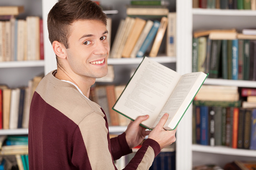 Cheerful young man holding a book and looking over shoulder while standing at the library