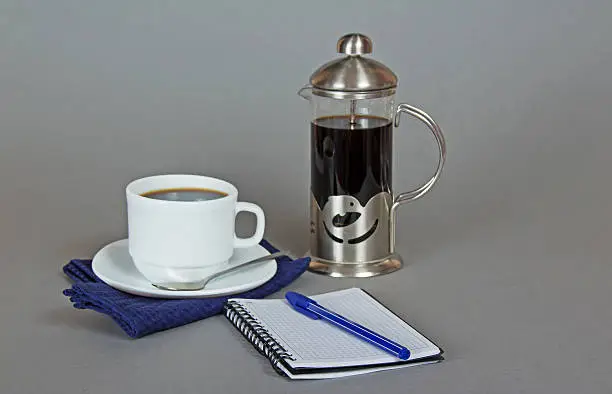Coffee maker, cup of coffee on a napkin, a sketchpad and a ballpoint on a gray background
