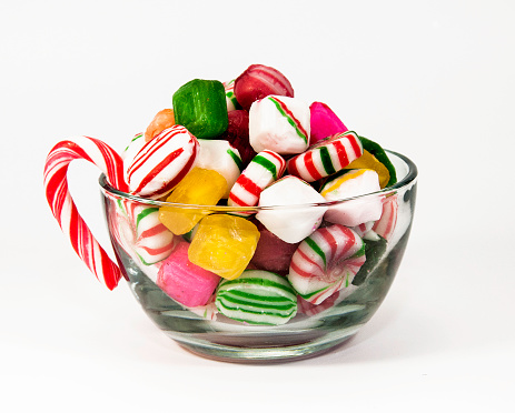 Glass bowl with candy cane handle filled with multicolored hard Christmas candies against a white background