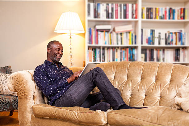 sorridente uomo maturo con tavoletta digitale nel salotto - reading book men sofa foto e immagini stock