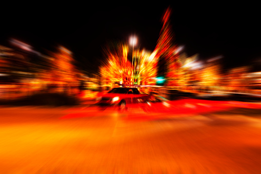 Defocused, zoomed image of a downtown city street at night. Unique photographic effects create a blurred motion vanishing point. Red, orange and yellow hues with great copyspace at bottom. A red vehicle in center of image driving down the busy road. Bright lights in background. Taillights, headlights.