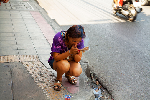 Bangkok, Thailand - June 2nd, 2013: A Thai woman is praying with incense sticks o sidewalk in street of Bangkok. Woman has placed some food, kind of dessert on sidewalk and is praying. Sticks are burning. At right side is ruling traffic in street.