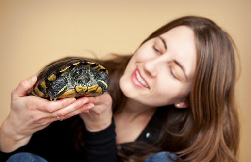 A selective focused image of a woman smiling and looking at the red eared slider turtle that she is holding.  Focus is on the turtle's face only.