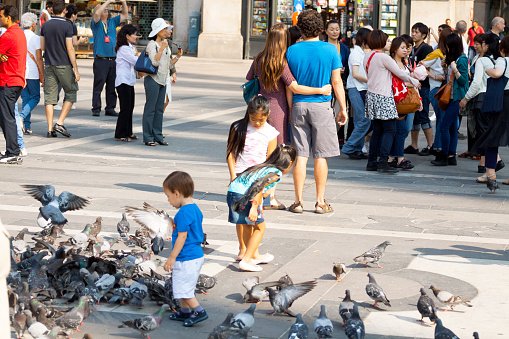Milan, Italy - September, 1st 2013: Many tourists are standing on square Piazza Duomo in summer. In forgeround are some children playing with pidgeons. Focus is on couple watching entrance of Galleria Vittorio Emanuele II in background.
