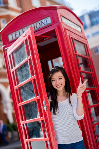 Happy Asian tourist in London in a telephone booth making a peace sign and looking at the camera