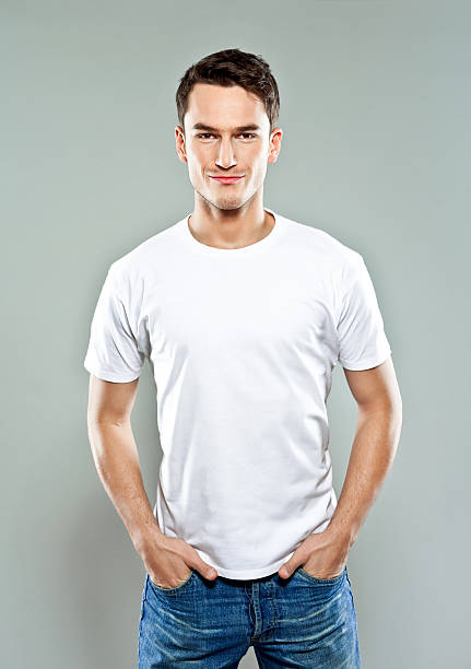 Cheerful young man Portrait of confident young man wearing white t-shirt, standing with hands in pockets and smiling. Studio shot, grey background. hands in pockets stock pictures, royalty-free photos & images