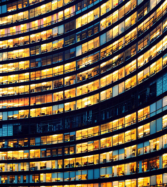 Office building at night with illuminated windows The image depicts workers working late in an office at night. The employees are seen as silhouettes against their lit offices that have large windows. The building is lit by the "blue hour" evening sky. The building and its windows cover the entire canvas of the image. It is apparent that the building may have some sort of curvature to the architecture. While most of the building reflects patterns of yellow light, a small portion of it reflects the blue light. There is no border around the image. The windows on the edges and corners of the image are truncated in the view. bustling city stock pictures, royalty-free photos & images
