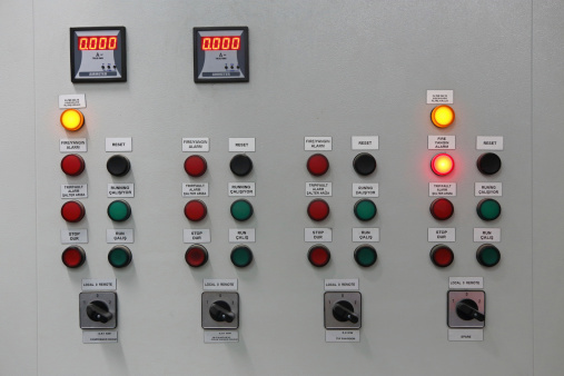 A close up view from an electrical control panel that displays digital amper gauges, control lamps, and switches. Some lamps on the row allocated for fire alarm system are lit.