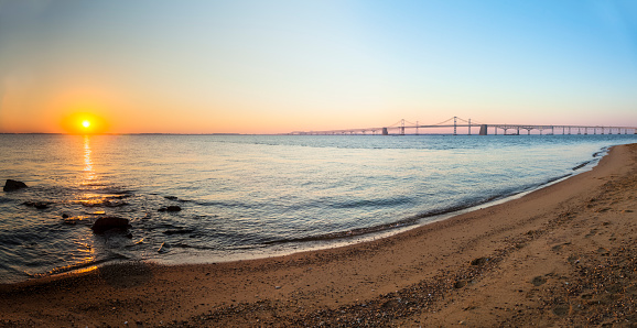 Ripples of the Chesapeake Bay gently lap the golden sands of Sandy Point State Park as the Chesapeake Bay Bridge stretches out along the horizon as the sun slowly rises.
