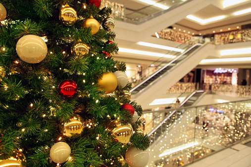 Close up of a Christmas tree with decorations and lights, ornating a modern shopping mall main space, out of focus, with lights, stairs and escalators on the background, modern architecture