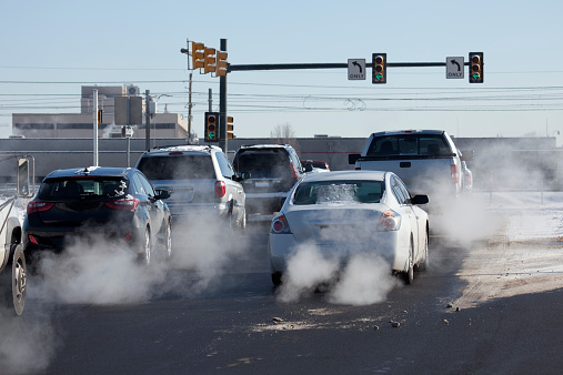 At a busy intersection with traffic signals and stop lights in Denver, Colorado on a winter day, exhaust pours out of tailpipes from accelerating cars, trucks, and other vehicles as they drive onto a snowy Santa Fe Drive when the left arrow turns green.