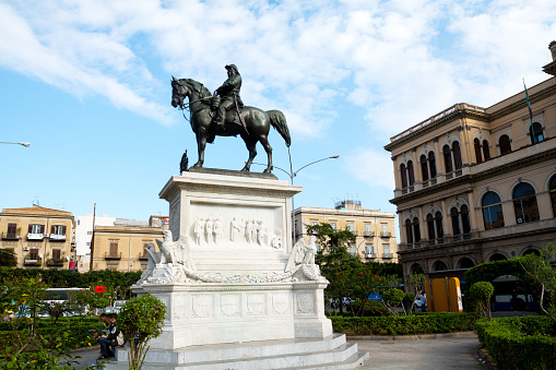 Palermo, Italy - November, 26th 2011: Statue of Vittorio Emanuele II on square Piazza Giulio Cesare with station at right side. At left bottom of statue are sitting three men. Equestrian statue was made by Civiletti in 1887.