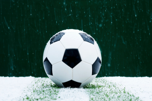One black and white soccer ball has been left out in the snow. There is a slight dusting of snow accumulating on the soccer ball but there is a lot of snow accumulating on the grass. The grass is almost completely covered in snow with only a few blades visible. The snow is still falling and it appears to be night.