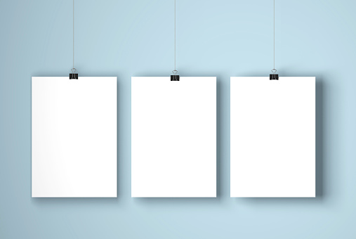 A blank white posters suspended against a plain blue wall. The paper is being held by a paper clip which is hanging on a piece of string. Light is coming from the left of the image which casts a soft shadow onto the background. The paper is in portrait orientation and is ready for you to add your own designs and artwork.