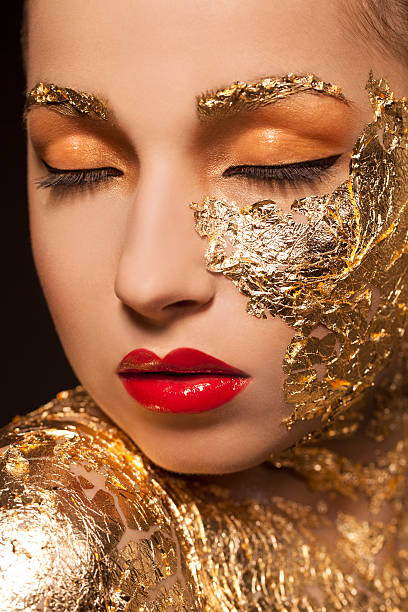 A dramatic artistic fashion portrait of a woman with shimmering gold accents on her face and nails and black paint on her body, set against a black background emphasizing elegance and mystery.